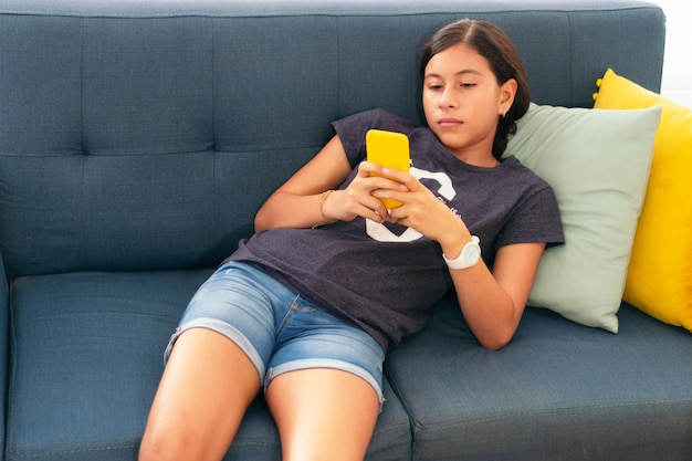Étudiante hispanique assise sur son canapé à la maison à l'aide d'un smartphone.