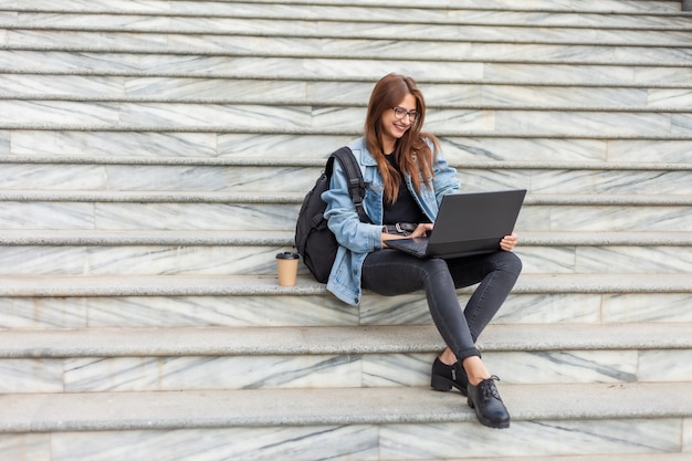 Étudiante jeune femme moderne dans une veste en jean assis sur les escaliers avec ordinateur portable. Apprentissage à distance. Concept de jeunesse moderne.