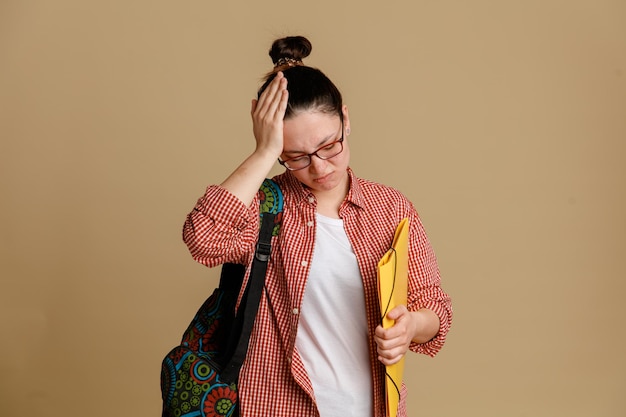 Étudiante jeune femme en vêtements décontractés portant des lunettes avec sac à dos tenant le dossier à la confusion avec la main sur la tête pour une grosse erreur debout sur fond marron