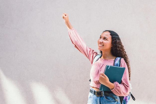 Étudiante latine souriante levant son bras en signe de célébration devant un mur. Une saine confiance en soi