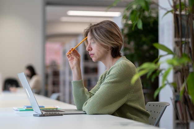 Étudiante mature concentrée étudiant en ligne sur un ordinateur portable dans une bibliothèque moderne entourée de verdure