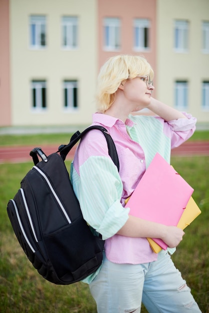 Étudiante pensive tenant un dossier rose avec des livres et portant un sac d'école marchant sur le campus de l'école