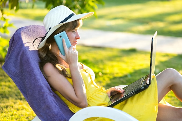 Étudiante en robe d'été jaune reposant sur une pelouse verte dans un parc d'été étudiant sur un ordinateur portable ayant une conversation sur un téléphone portable. Faire des affaires et apprendre pendant le concept de quarantaine.