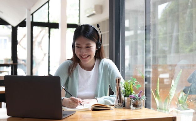 Étudiante souriante portant un casque sans fil étude en ligne avec un professeur skype heureuse jeune femme apprendre la langue écouter conférence regarder webinaire écrire des notes regarder un ordinateur portable s'asseoir dans un café éducation à distancexA