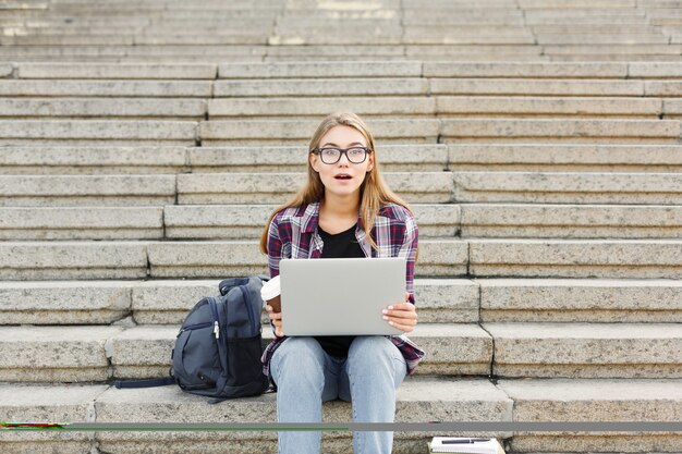 Étudiante surprise assise dans les escaliers travaillant avec un ordinateur portable, se préparant aux examens et buvant du café à l'extérieur, sur le campus universitaire. Concept de technologie, d'éducation et de travail à distance, espace de copie
