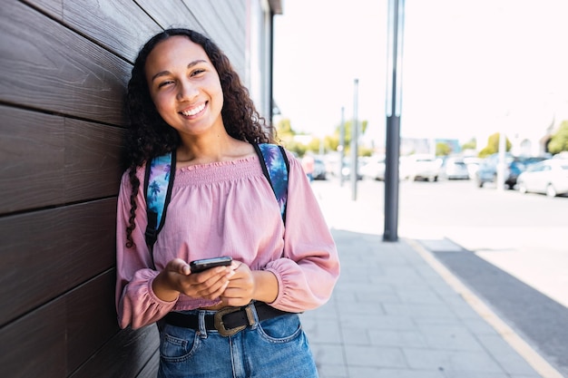 Étudiante universitaire hispanique souriante debout et tenant un téléphone portable sur le trottoir.