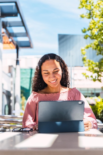 Étudiante universitaire latine souriante et utilisant une tablette assise à l'extérieur du centre commercial pendant son temps libre