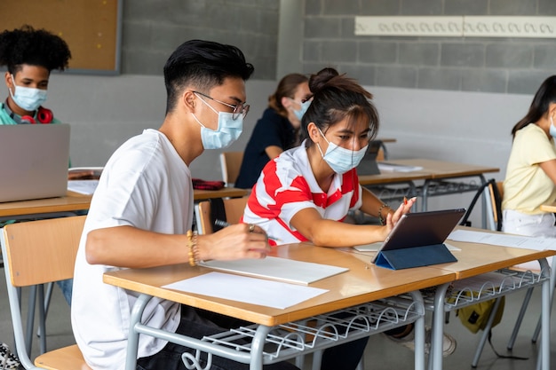 Étudiants adolescents multiraciaux avec masque facial à l'aide d'une tablette en classe