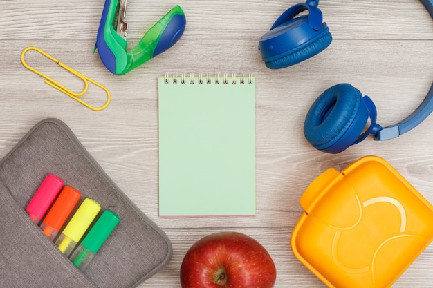 Étui à crayons avec feutres de couleur et marqueur, pomme, cahier vert, agrafeuse, écouteurs et boîte à lunch sur fond de bois gris. Vue de dessus avec espace de copie. Retour au concept de l'école. Fournitures scolaires.