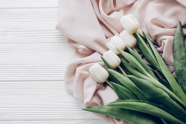 Tulipes blanches élégantes sur tissu doux beige et vue de dessus de fond de table en bois rustique bonjour printemps plat poser douceur lumière tendresse moment atmosphérique espace pour texte image moderne