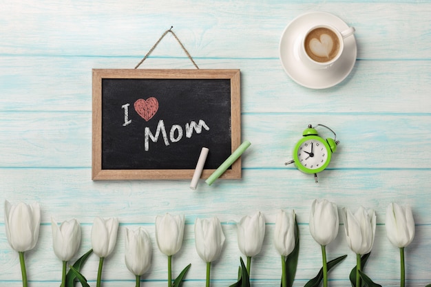 Tulipes blanches avec un tableau noir, une tasse de café et un réveil sur des planches en bois bleus. Fête des mères