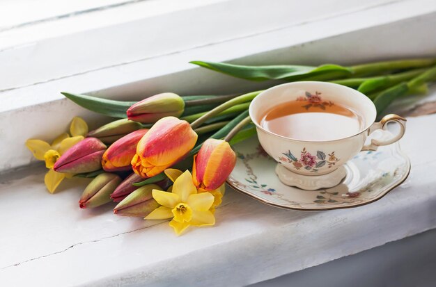 Tulipes, jonquilles et une tasse de thé sur le rebord de la fenêtre en bois. Mise au point sélective.