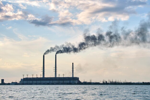 Tuyaux hauts de centrale électrique au charbon avec fumée noire se déplaçant vers le haut, atmosphère polluante au-dessus de l'eau du lac.