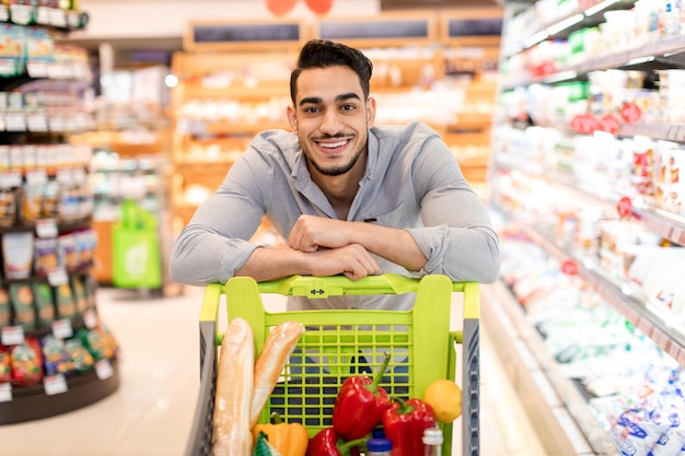 Type arabe posant avec le chariot faisant des emplettes dans le supermarché moderne