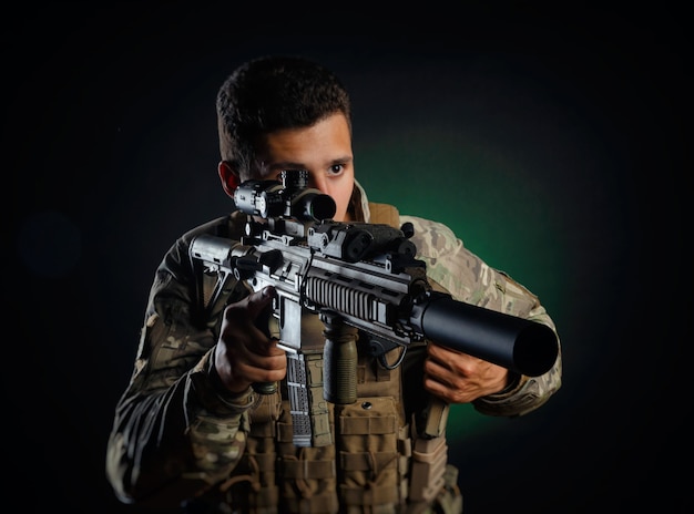 Un type brutal dans des combinaisons d'airsoft militaires pose avec une arme dans ses mains sur un fond foncé