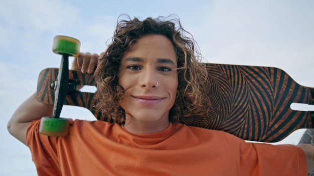 Un type joyeux et joyeux souriant à la caméra tenant un skateboard sur les épaules en gros plan