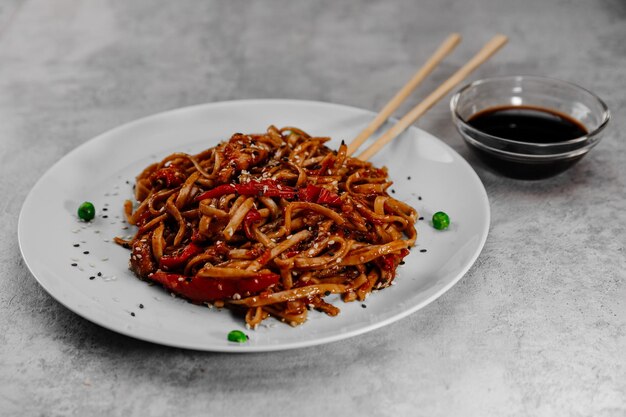 Udon épicé au poulet sur une assiette blanche avec de la sauce soja dans un bol en verre sur fond gris. Côté