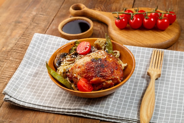 Udon avec légumes et poulet sur une table dans une assiette en bois sur une serviette à côté de tomates et une fourchette en bois sur une planche