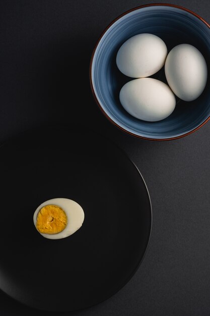 Œuf à la coque avec du jaune d'oeuf dans une assiette noire près d'un bol avec des œufs blancs sur une surface minimale sombre sombre de mauvaise humeur, vue d'angle, joyeuses Pâques