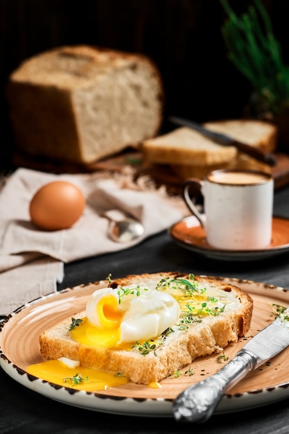 Œuf à la coque (poché) sur une tranche de pain recouverte de crème au beurre et d'herbes, sur plaque d'argile sur table en bois noir