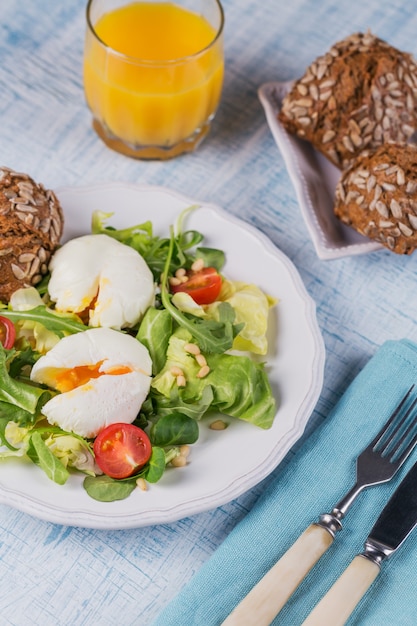 Œuf poché avec salade verte, tomates, pain complet et jus d'orange sur bois bleu.