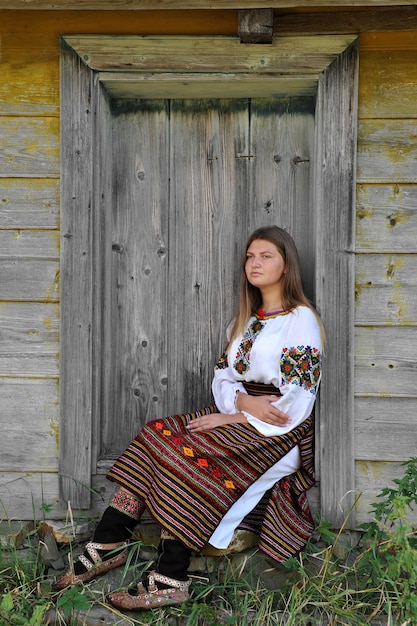 Une Ukrainienne en vêtements hutsul nationaux est assise sur le seuil d'une vieille maison en bois