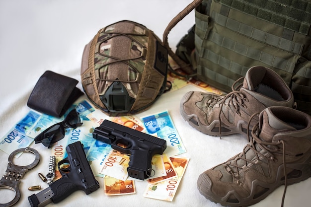 Uniforme et équipement militaire, munitions. Armure corporelle, armes à feu, casque pare-balles, menottes, lunettes de soleil et bottes militaires avec des billets de banque en nouveaux shekels israéliens. Armes pour airsoft et manifestations urbaines