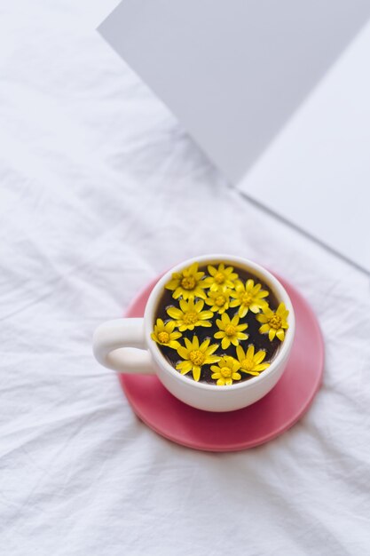 Ð¡up avec des fleurs jaunes à l'intérieur, sur un lit blanc le matin avec bloc-notes