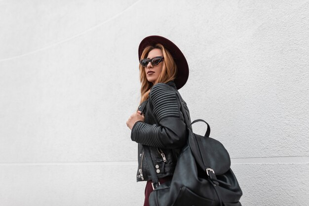 Urban jolie jeune femme hipster dans des lunettes de soleil sombres à la mode dans un chapeau chic dans une veste en cuir avec un sac à dos élégant noir