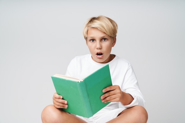 ute excité petit garçon blond portant un sweat-shirt debout isolé sur un mur gris, lisant un livre