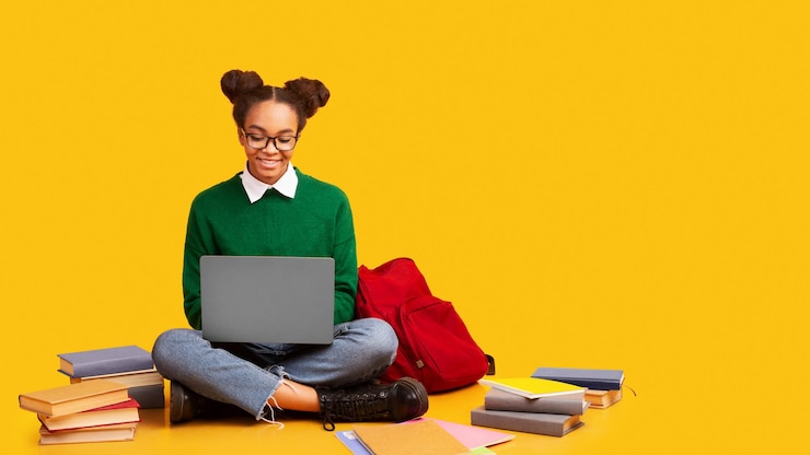 Photo utilisant un ordinateur portable une adolescente noire heureuse faisant ses devoirs assise sur le sol à l'arrière-plan jaune du studio