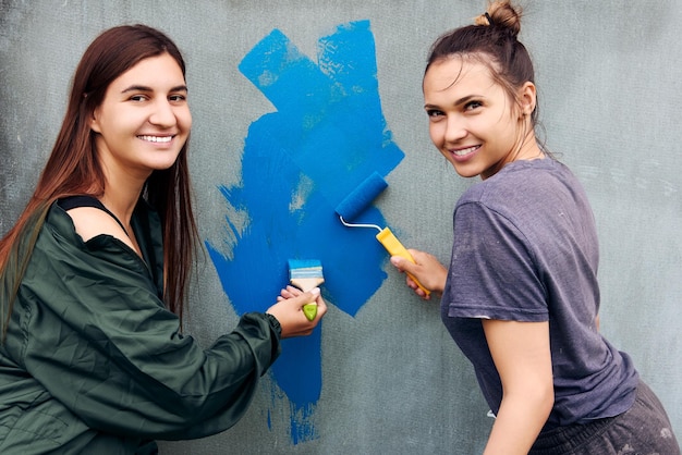 Utilisant de la peinture bleue pour peindre des murs gris, de jeunes femmes souriantes travaillant comme peintres de maisons.
