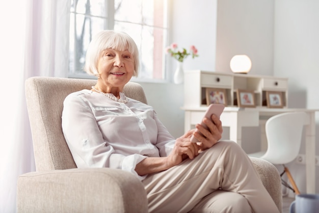 Utilisateur avancé. Cheerful senior woman assis dans un fauteuil et souriant tout en surfant sur Internet sur son téléphone