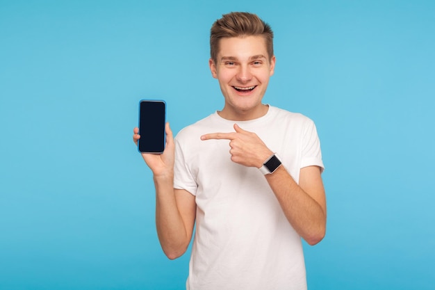 Utilisateur satisfait du téléphone portable Portrait d'un homme joyeux et joyeux en t-shirt blanc pointant vers un téléphone portable et regardant la caméra avec un sourire heureux et excité tourné en studio à l'intérieur isolé sur fond bleu