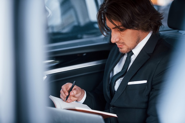 Utilisation du bloc-notes. Jeune homme d'affaires en costume noir et cravate à l'intérieur d'une automobile moderne.