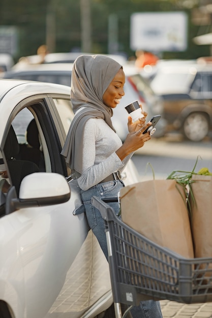 Utilisation du smartphone en attendant. Femme d'origine africaine sur la station de recharge de voitures électriques pendant la journée. Véhicule tout neuf.