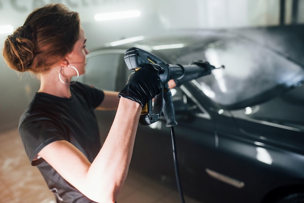 Utilisation d'équipement avec de l'eau à haute pression Une automobile noire moderne est nettoyée par une femme à l'intérieur de la station de lavage de voiture