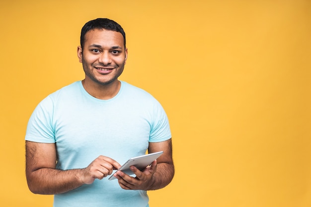 Utilisation de la tablette. Heureux gagnant ! Jeune bel homme indien afro-américain souriant tenant une tablette et jouant à des jeux ou utilisant une application de réservation isolée sur fond jaune.