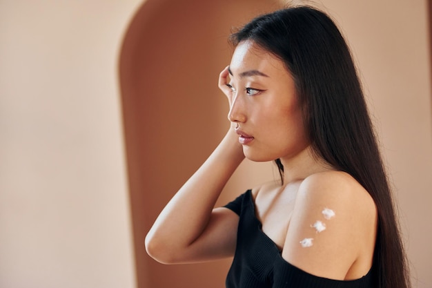 Utilise la crème de beauté Jeune femme asiatique sérieuse debout à l'intérieur