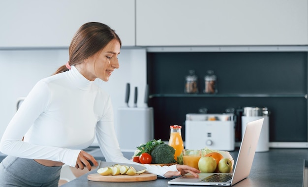 Utilise un ordinateur portable Jeune femme européenne est à l'intérieur à la cuisine à l'intérieur avec des aliments sains