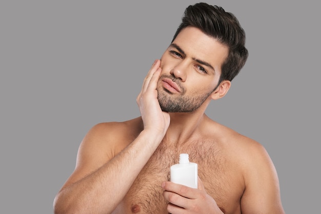 Utilisé pour avoir l'air parfait. Beau jeune homme appliquant une lotion après-rasage et souriant en se tenant debout sur fond gris