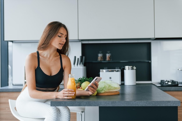 Utilise un smartphone Jeune femme européenne est à l'intérieur à la cuisine à l'intérieur avec des aliments sains