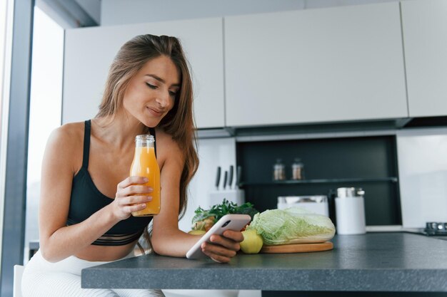 Utilise un smartphone Jeune femme européenne est à l'intérieur à la cuisine à l'intérieur avec des aliments sains