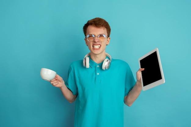 Utiliser des appareils, boire du café. Portrait moderne du jeune homme caucasien isolé sur mur bleu, monochrome. Beau modèle. Concept d'émotions humaines, expression faciale, ventes, publicité, tendance.