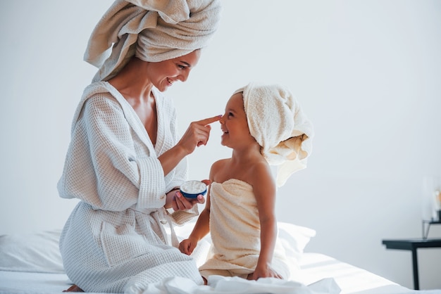 Utiliser de la crème pour nettoyer la peau. Une jeune mère avec sa fille a une journée de beauté à l'intérieur dans une salle blanche.