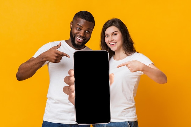 Utilisez cette nouvelle application heureux couple multiracial pointant vers un smartphone avec une maquette d'écran vide