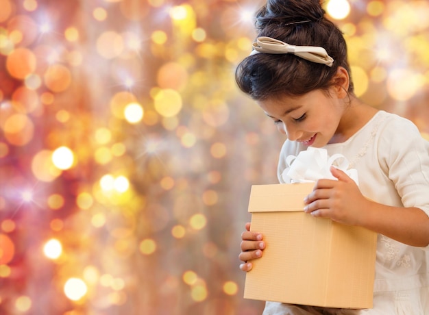 vacances, cadeaux, noël, enfance et concept de personnes - petite fille souriante avec boîte-cadeau sur fond de lumières