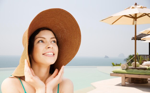 vacances d'été, les gens, le tourisme, les voyages et le concept de vacances - heureuse jeune femme au chapeau de paille sur la plage à l'arrière-plan de la station