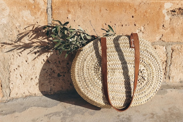 Photo vacances d'été nature morte panier rond en paille française avec poignées en cuir et feuilles et branches d'olivier vert au soleil fond de mur en grès doré concept de style de vie et de mode de voyage