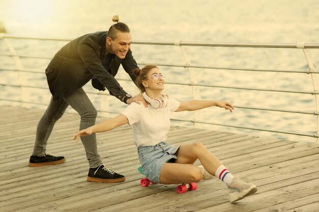 Vacances d'été pour les jeunes. jeune couple d'amoureux sur la promenade de la plage. Le gars pousse la fille assise sur une planche à roulettes. Notion de liberté